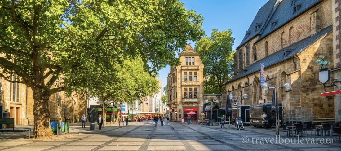 Een breed, zonnig stadsplein in het centrum van Dortmund met historische kerken, bomen en wandelende mensen.