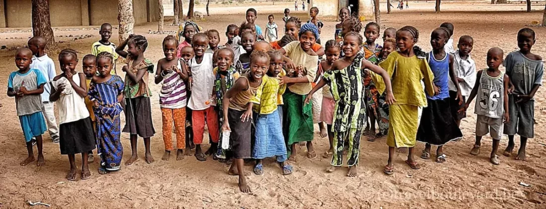 Een grote groep lachende kinderen die naar de camera rent op een zanderig schoolplein in Gambia.
