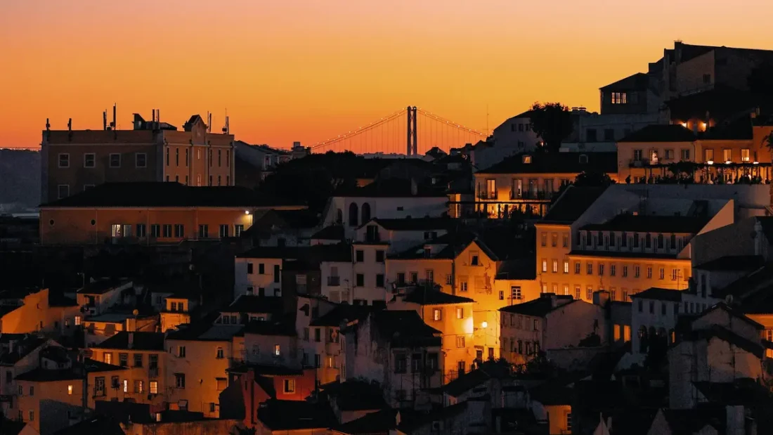 Panoramic view over the illuminated houses of Lisbon and the Ponte 25 de Abril bridge during sunset.