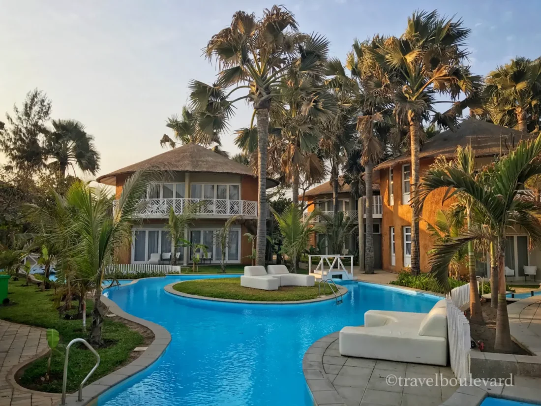 Uitzicht op het Balafon Beach Resort in Gambia met een blauw zwembad dat rondom traditionele kamers en palmbomen slingert.