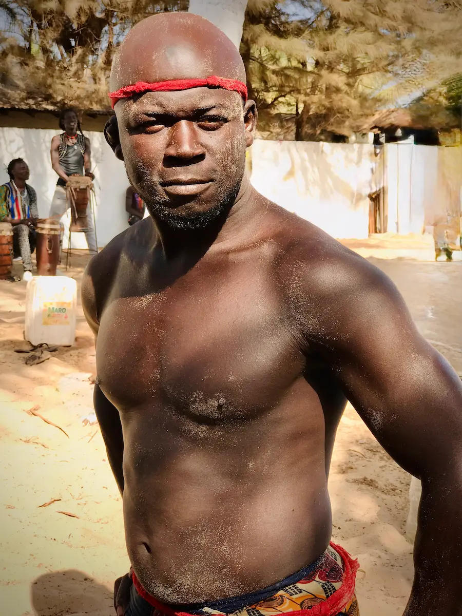 Een gespierde worstelaar met een rode hoofdband en zand op zijn lichaam staat in een arena in Gambia.