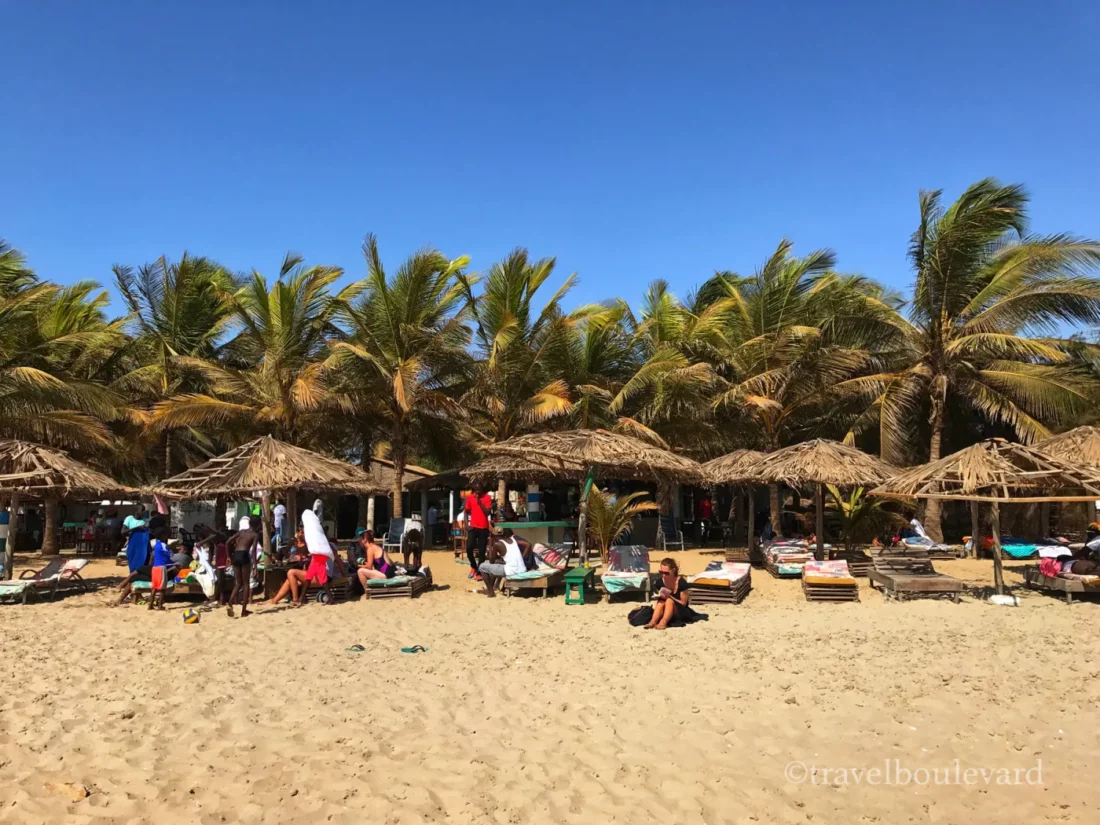 Een zonnig breed zandstrand in Gambia met rieten parasols, ligbedden, palmbomen en mensen die ontspannen bij een strandbar.