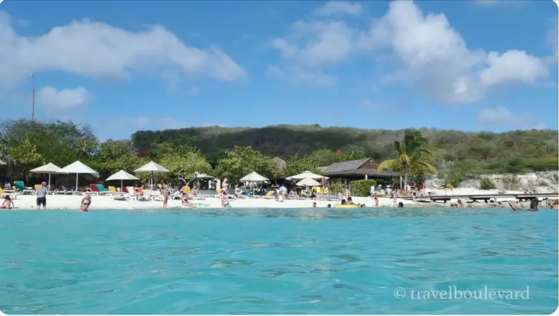 Zicht vanaf het turquoise water op het witte zandstrand van Porto Mari op Curaçao, met strandparasols en de houten pier.