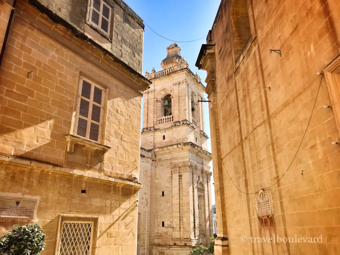 View through a narrow street of the St. Lawrence Church bell tower in Vittoriosa, Malta, with historic limestone buildings and a bright blue sky.