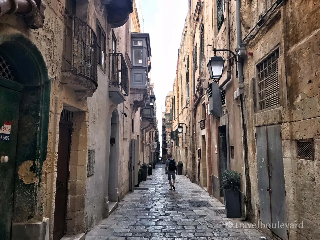 A narrow historic street in Valletta with limestone buildings, traditional blue wooden balconies, and lanterns on the facades.